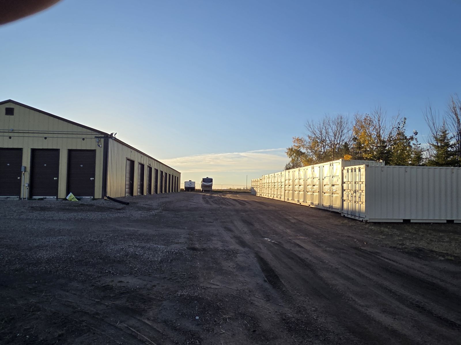 Wide gravel drive aisle between a storage unit building with roll-up doors and a row of white seacans